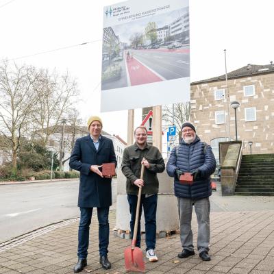 Drei Männer stehen mit einem Spaten vor einem großen Bauschild.