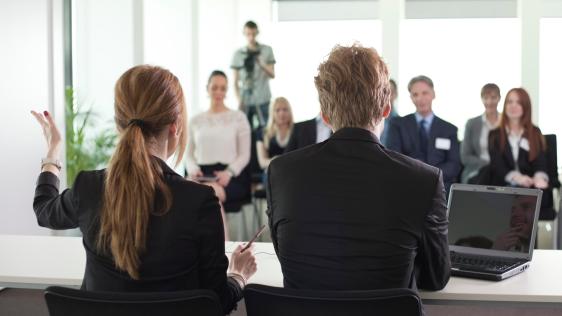 Mann und Frau sitzen an einem Tisch, während eine Gruppe von Menschen ihnen gegenüber sitzt.