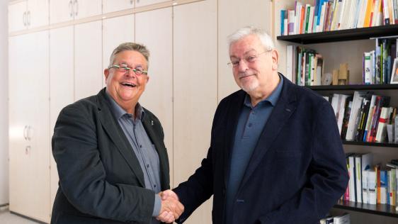 Dr. Daniela Grobe begrüßt Wolfgang Wenner (Ombudsperson der Stadt Mülheim an der Ruhr). Im Hintergrund ist eine Schrankwand und ein Bücherregal.
