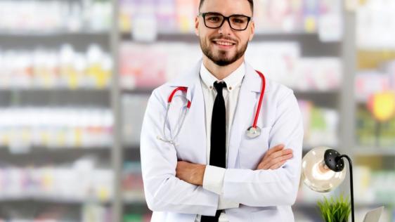 Ein Apotheker mit schwarzer Brille, freundlichem Blick und verschränkten Armen steht in seiner Apotheke. Neben ihm steht eine schwarze Schreibtischlampe und eine grüne Topfpfanze.