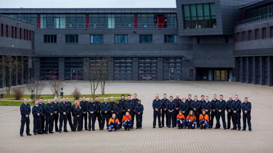 Auf dem Foto sieht man eine größere Gruppe von Mitgliedern der Freiwilligen Feuerwehr Mülheim an der Ruhr, die geschlossen vor einem modernen Gebäudekomplex – vermutlich einer Feuerwache – in mehreren Reihen posieren. Einige Personen tragen die typische dunkelblaue Einsatzkleidung der Feuerwehr, während ein paar jüngere Mitglieder orangefarbene Jugendfeuerwehr-Bekleidung tragen.