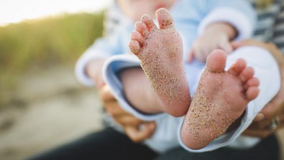 Person hält ein neugeborenes Baby auf dem Arm, der Fokus steht auf die nackten Füßchen, an denen Sand haftet. Standesamt, Anmeldung einer Geburt, Adoption, Pflegeeltern, Familie, Kindertagespflege