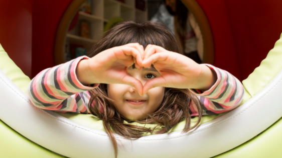 Kleines Mädchen liegt in einer Spielhöhle und formt mit ihren Händen ein Herz. Kindertagespflege