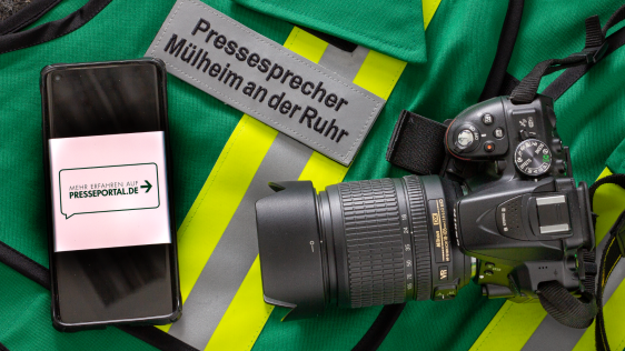 Auf einer grünen Warnjacke mit Reflexstreifen liegen ein graues Namensschild „Pressesprecher Mülheim an der Ruhr“, ein Smartphone mit der Anzeige „Mehr erfahren auf presseportal.de“ und eine DSLR‑Kamera mit Zoomobjektiv.