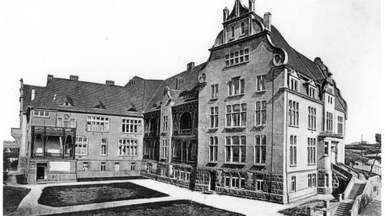 Das Bild zeigt ein mehrstöckiges Gebäude aus dem Jahre 1907. Vor dem Gebäude befindet sich eine angelegte Grünanlage.