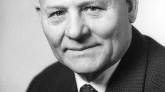 Auf dem Portraitfoto ist ein älterer, lächelnder Herr ohne Haare zu sehen. Er trägt eine schwarze Anzugjacke, ein weißes Hemd und eine schwarze Krawatte.