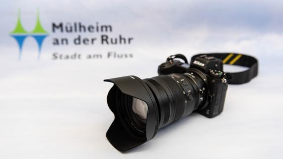 Schwarze Nikon Kamera mit Objektiv und Gegenlichtblende auf weißem Untergrund, im Hintergrund unscharf das Logo von Mülheim an der Ruhr mit dem Text 'Stadt am Fluss'
