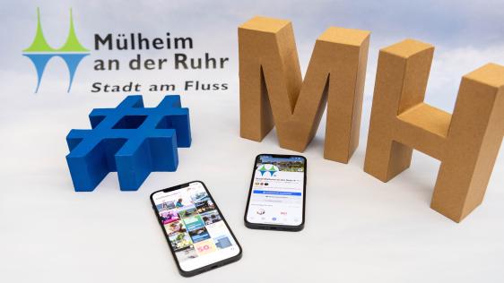 Großes blaues Hashtag-Symbol, große Buchstaben 'MH' aus Karton, zwei Smartphones mit Social-Media-Seiten der Stadt Mülheim an der Ruhr