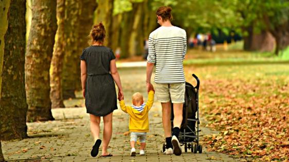 Zwei Personen die in der Mitte ein Kleinkind an den Händen halten. Eine Person schiebt dabei noch einen Kinderwagen