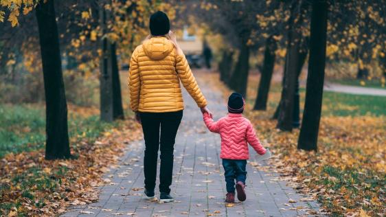 Frau mit kleiner Tochter an der Hand beim Spaziergang im Park, Laub liegt auf dem Weg.