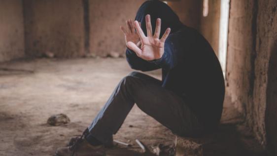 Ein Jugendlicher in einem schwarzen Hoodie mit Kapuze über dem Kopf sitzt in einem verlassenen Gebäude und zeigt eine Abwehrhaltung mit seinen Händen.
