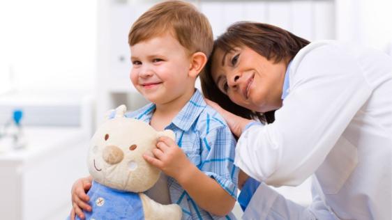 Ärztin untersucht einen kleinen Jungen mit einem Teddy auf dem Arm in der Kinderarztpraxis