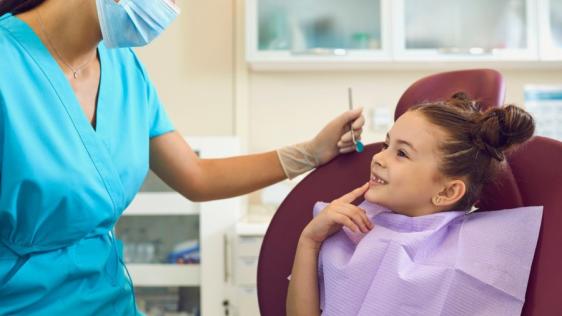 Ein junges Mädchen mit braunen Haaren und zwei Zöpfen sitzt lächelnd in einem Behandlungsstuhl in einer Praxis für Zahnmedizin. Gegenüber sitzt eine Zahnärztin in einer blauen Praxisbekleidung und hält in der linken Hand einen Mundspiegel.