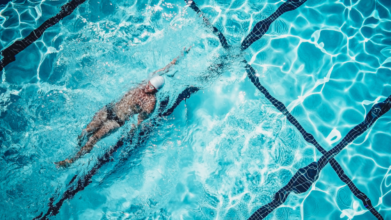 Eine Person schwimmt in einem Schwimmbecken im Rückenschwimmstil. Das klare blaue Wasser ist von schwarzen Bahnmarkierungen durchzogen, und Sonnenlicht reflektiert auf der Wasseroberfläche.