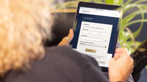 Eine Person hält ein Tablet in der Hand und füllt online einen Fragebogen zum Zensus aus. Der Bildschirm zeigt ein Eingabeformular. Im Hintergrund ist eine Pflanze zu sehen.
