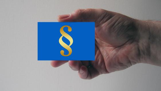 Ein Mensch hält ein blaues, kleines Schild in der Hand, auf dem Bild ist ein goldenes Paragraphen Zeichen zu sehen
