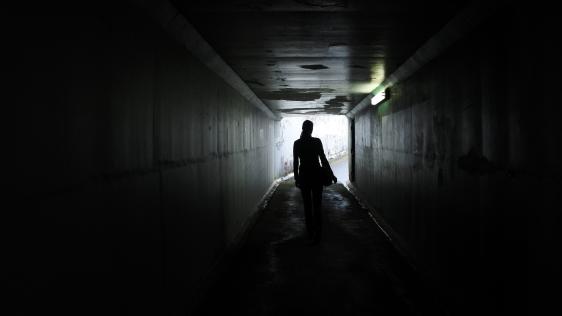 Dunkle Silhouette einer Frau, die durch einen dunklen Tunnel läuft. Übergriffe auf Frauen und Mädchen finden alltäglich und öffentlich statt.