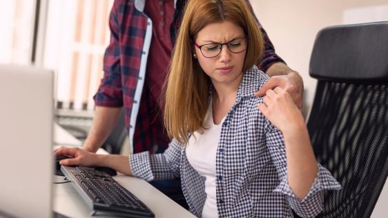 Bild zeigt eine junge Frau am PC-Arbeitsplatz, ein Kollege stützt sich auf ihrem Schreibtisch ab und hat einen Arm um sie gelegt, den sie verägert wegschiebt.