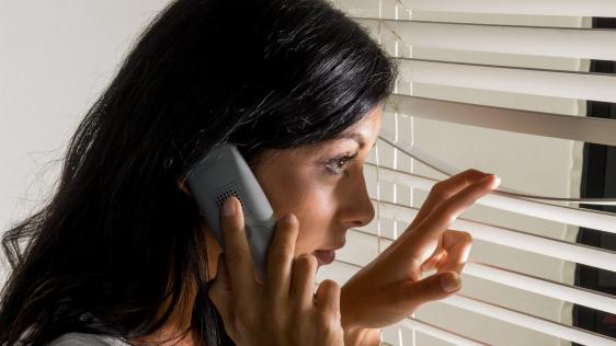 Junge Frau mit Telefon am Ohr schaut besorgt durch die Rollos an ihrem Fenster. Stalking-Handlungen beeinträchtigen den Tagesablauf und die Lebensqualität der Opfer erheblich.