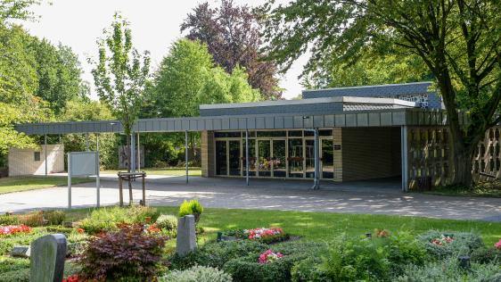 Trauerhalle von außen in Heißen. Im Vordergrund Urnengräber mit Grabmalen. Die Trauerhalle liegt im Schatten der umstehenden Bäume