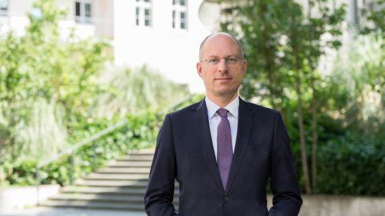 Mann in dunklem Anzug und weißem Hemd steht vor Treppe und begrüntem Hintergrund