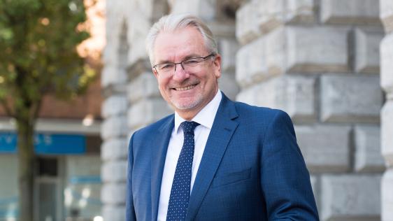 Mann mit grauen Haaren trägt blauen Anzug, weiße Hemd und dunkelblaue Krawatte, steht vor dem Rathaus Mülheim
