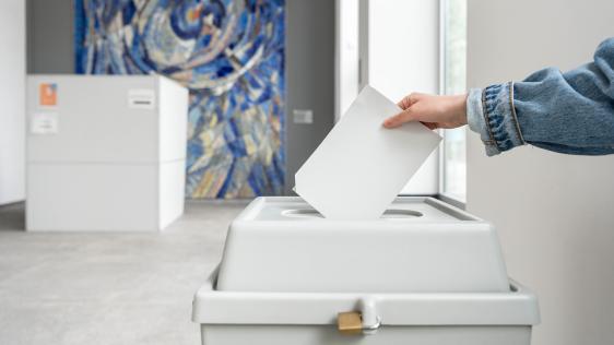 Hand wirft Stimmzettel in Wahlurne bei Europawahlen in modernem Wahllokal