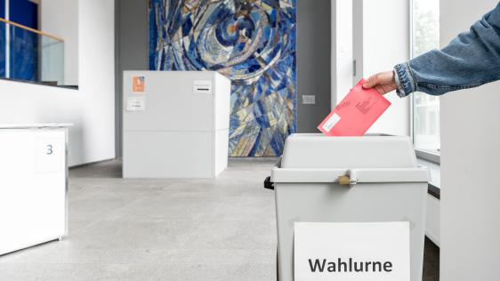 Person wirft roten Wahlzettel in weiße Wahlurne mit Aufschrift 'Wahlurne' in hellem Raum mit abstraktem Wandbild