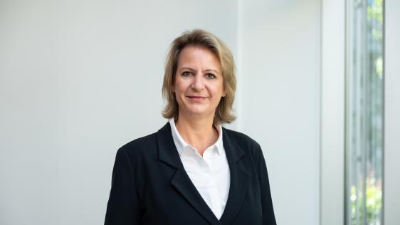 Frau mit schulterlangem blonden Haar trägt weißen Hemdkragen und schwarzen Blazer vor neutralem Hintergrund