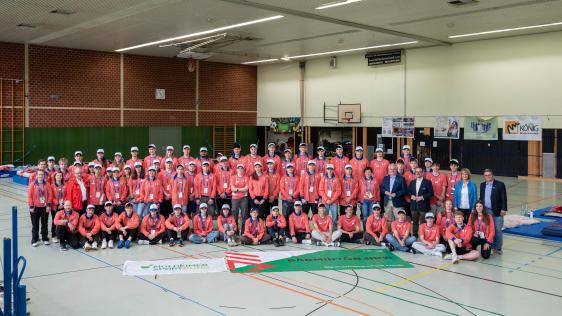 Große Gruppe von Jugendlichen und Erwachsenen in Sportkleidung und roten Jacken in der Sporthalle Von-der-Tann-Straße, Gruppenfoto beim FISU Badminton Youth Camp