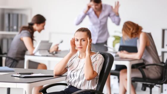 Eine Frau sitzt inmitten eines lauten Büros und hält sich den Kopf. Im Hintergrund sind drei weitere Frauen zu erkennen.