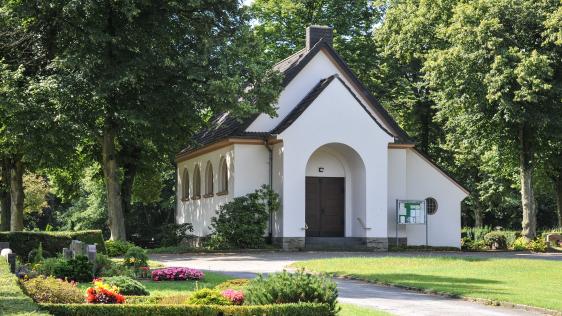 Trauerhalle in Broich von schräg links außen. Im Vordergrund sind Grabstätte zu erkennen. Die weiße Trauerhalle wird von den umstehenden Bäumen beschattet