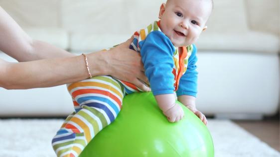 Ein lachendes Baby in einem bunten Strampelanzug sitzt auf einem grünen Gymnastikball und wird von dessen Mutter festgehalten 