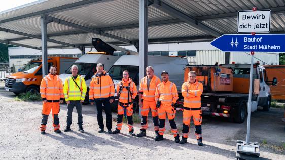 Mitarbeitende des Bauhofs in oranger und gelber Arbeitskleidung stehen vor einem Unterstand mit ihren Fahrzeugen. Neben ihnen steht eine blaues Richtungsschild mit der Aufschrift Bauhof Stadt Mülheim. Darüber ist ein weißes Schild mit dem Satz "Bewirb dich jetzt" montiert.