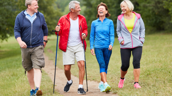Vier Senior*innen gehen auf einem Wald- und Wiesenweg spazieren. Die beiden Damen tragen sportliche Outfits. Einer der Männer hat Walking-Stöcke dabei. Die Gruppe ist guter Stimmung und hat sichtlich Spaß.