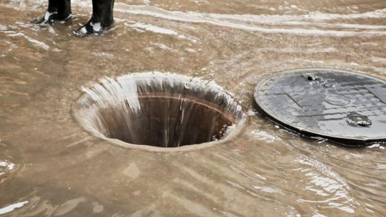 Ein Abwasserkanal wird dargestellt, in welchen literweise Wasser strömt. Im Hintergrund sind die schwarzen Gummistiefel einer Person zu erkennen.