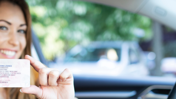 Eine Frau sitzt in einem Auto und zeigt mit einem Lächeln ihren Führerschein