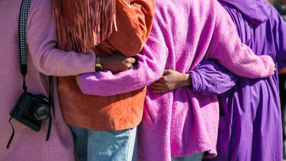 Von hinten fotografiert: Vier Frauen in knallbunten Mänteln halten sich in den Armen, Arm in Arm. Symbol für Gleichstellungsstelle, Zusammenhalt