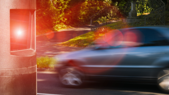 Eine Geschwindigkeitsmessstelle erfasst ein zu schnell fahrendes graues Auto auf der Straße.