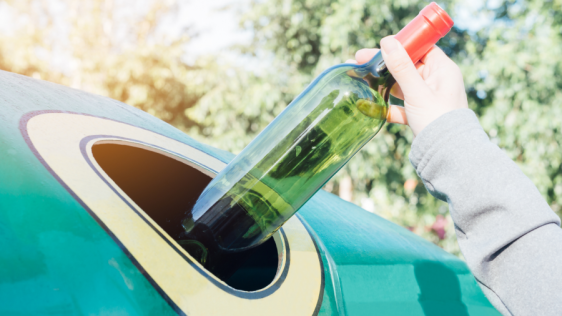 Hand einer Person, die eine leere Flasche aus grünem Glas in einen Glascontainer wirft.
