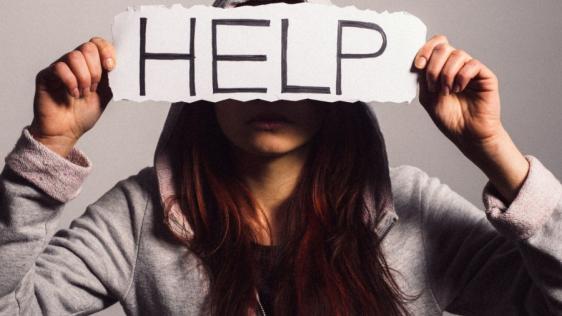 Eine junge Frau mit einem grauen Hoodie und langen braunen Haaren hält einen Zettel mit dem Wort Help vor ihr Gesicht.