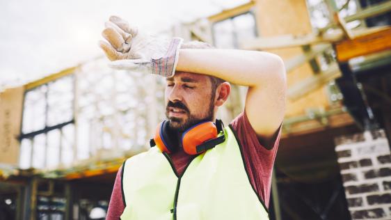 Ein Mann in Arbeitskleidung vor einer Industriekulisse hält sich die Hand vor die Stirn.