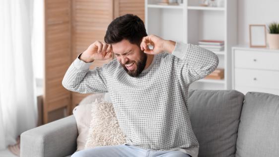 Ein Mann sitzt auf einem Sofa und hält sich die Ohren zu.