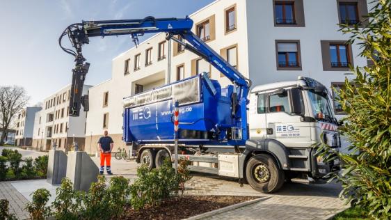 Leerung eines Unterflurbehälters mit einem großen Müllfahrzeug, dessen Kran ausgefahren ist, ein Mitarbeiter steht daneben.