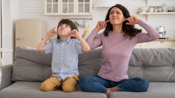 Kind und Frau sitzen auf einem Sofa, den Blick gen Decke gerichtet und halten sich jeweils die Ohren zu.