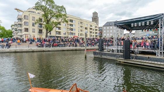 Blick vom Wasser aus in den Ruhrbania-Hafen anlässlich der offiziellen Eröffnung im Jahr 2014. Eine Bühne ist aufgebaut, Menschen stehen am Ufer. Im Hintergrund Bebauung und der Rathausturm.