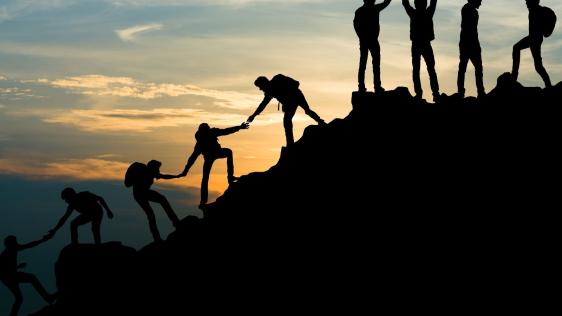 Eine Gruppe von Menschen besteigt einen Berg. Sie helfen sich untereinander die Bergspitze zu erreichen.