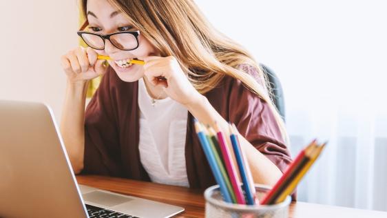 Junge Studierende sitzt vor aufgeklapptem Laptop, schaut auf den Bildschirm und beißt auf einen Stift