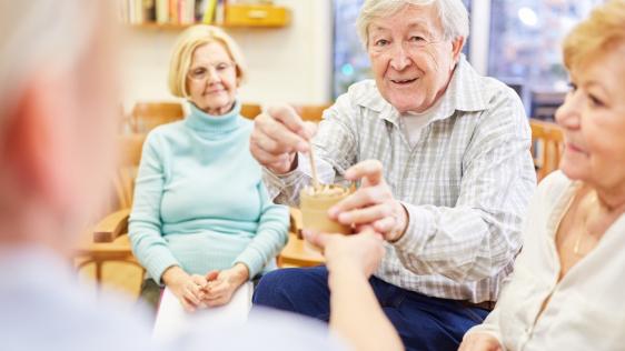 Auf dem Foto ist eine Gruppe von Seniorinnen und Senioren in einem Lehrraum zu sehen. Ein älterer Mann nimmt dabei von seinem Gegenüber (nur als Hinteransicht) ein Getränk entgegen.