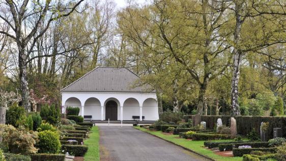 Die alte Trauerhalle des Hauptfriedhofs. Entlang eines Weges befinden sich links und rechts Grabstätten. Im Hintergrund ist die Trauerhalle zu sehen.